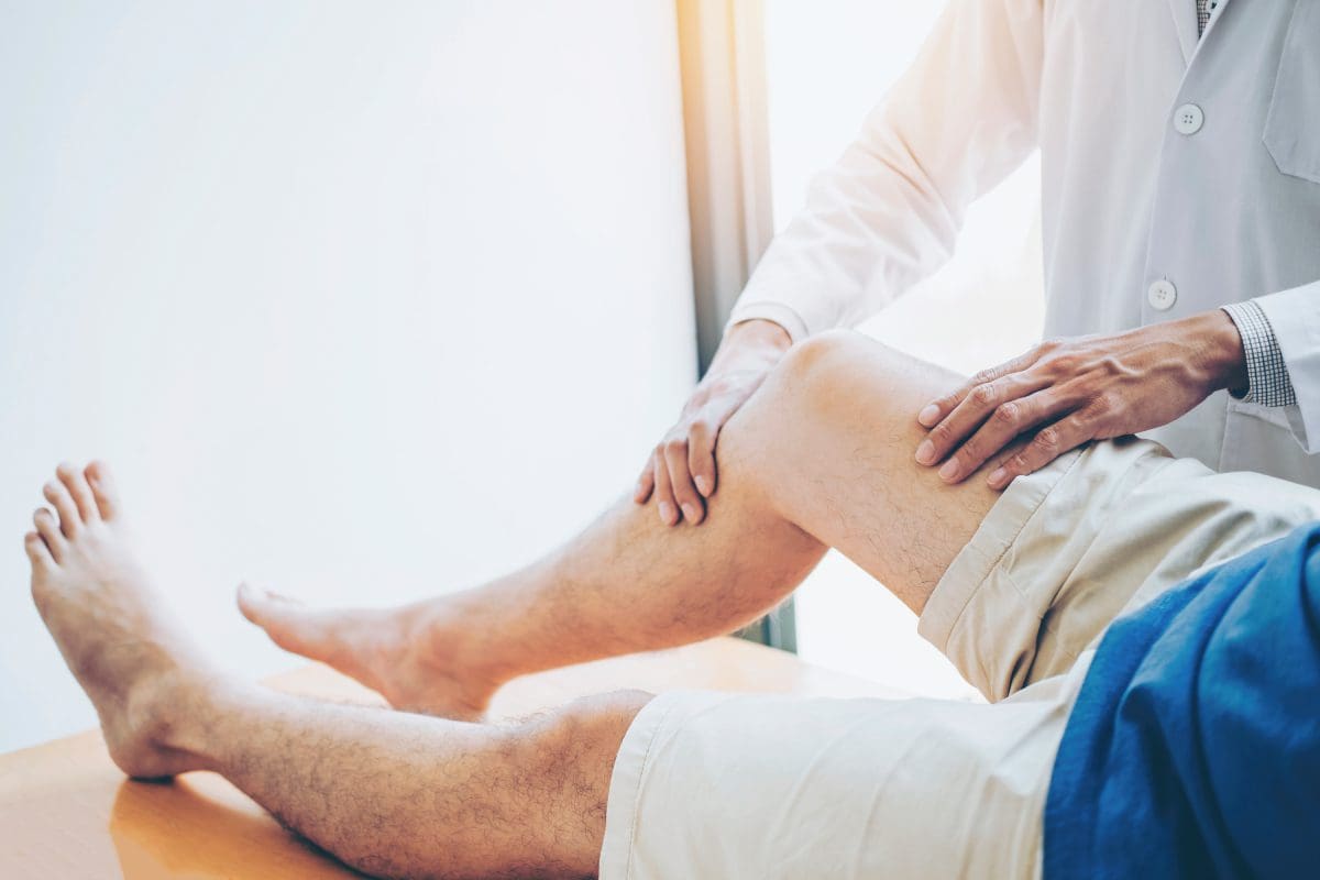 A person in a white coat examines a patient's knee while the patient lies on a table, wearing shorts.