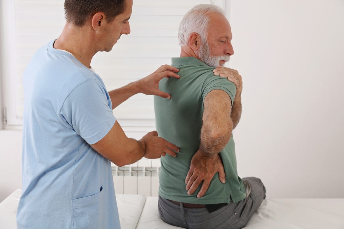 A physical therapist assists an elderly man during a back exercise in a medical setting. The man is twisting his upper body while sitting.