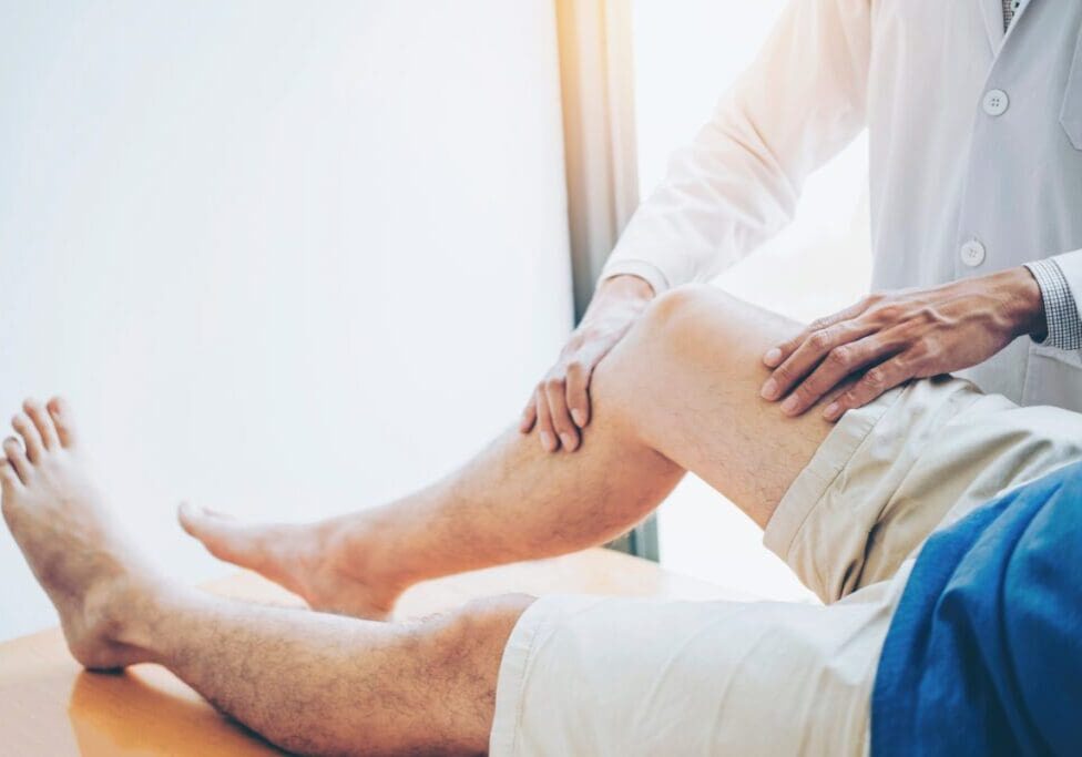 A person in a white coat examines a patient's knee while the patient lies on a table, wearing shorts.