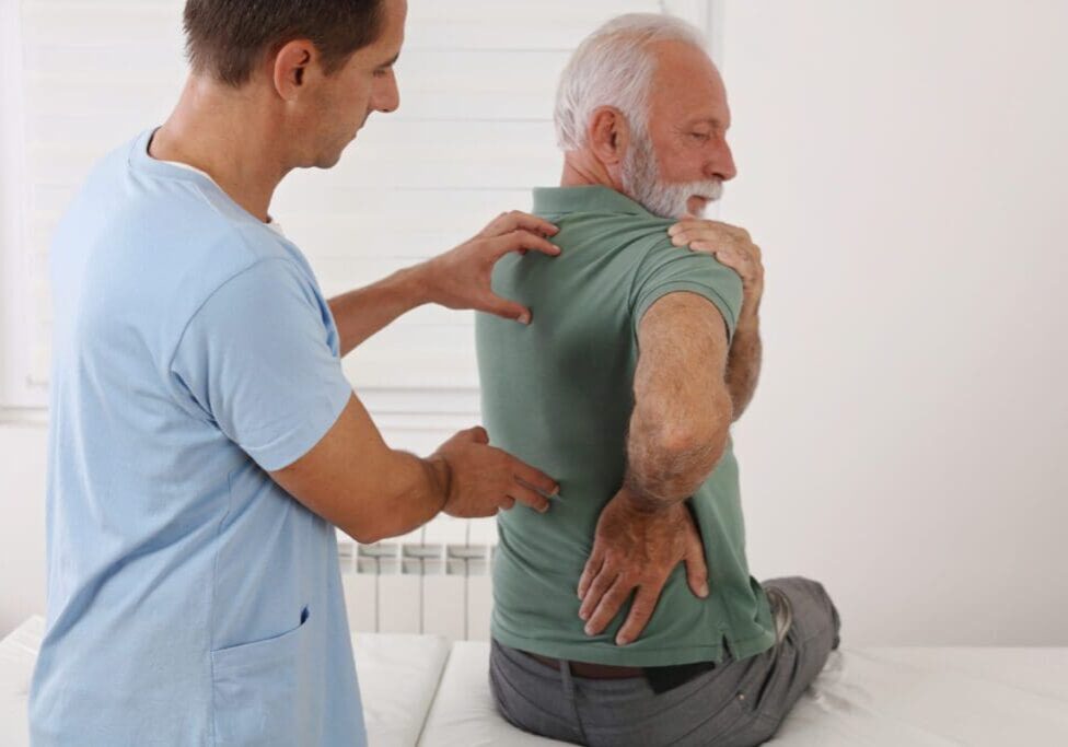 A physical therapist assists an elderly man during a back exercise in a medical setting. The man is twisting his upper body while sitting.